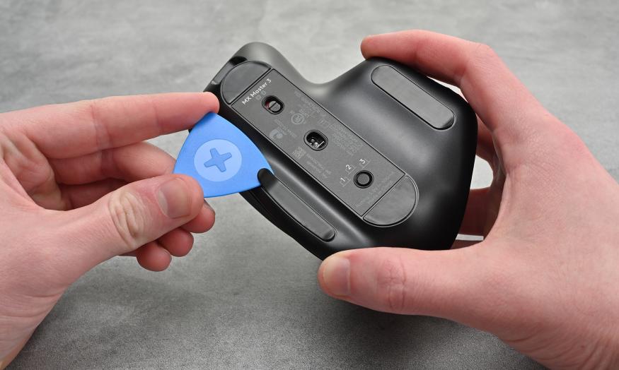 Photo of a person holding the Logitech MX Master 3 mouse with a blue spudger in the other hand pulling open a tab to open the device.