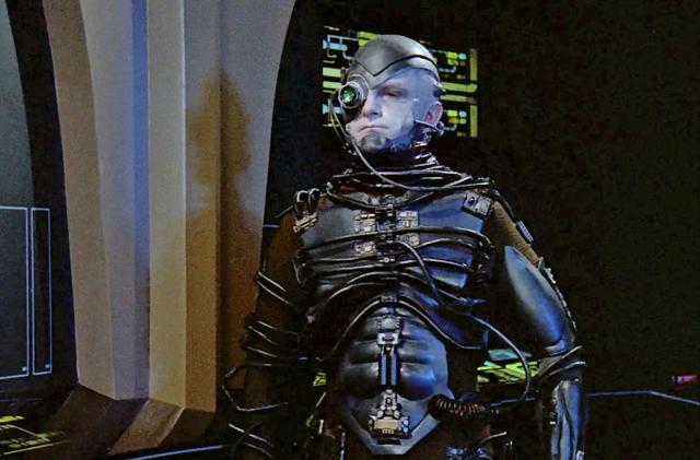 The Borg from Star Trek. A human / machine cyborg stands in a futuristic setting on a spaceship.  