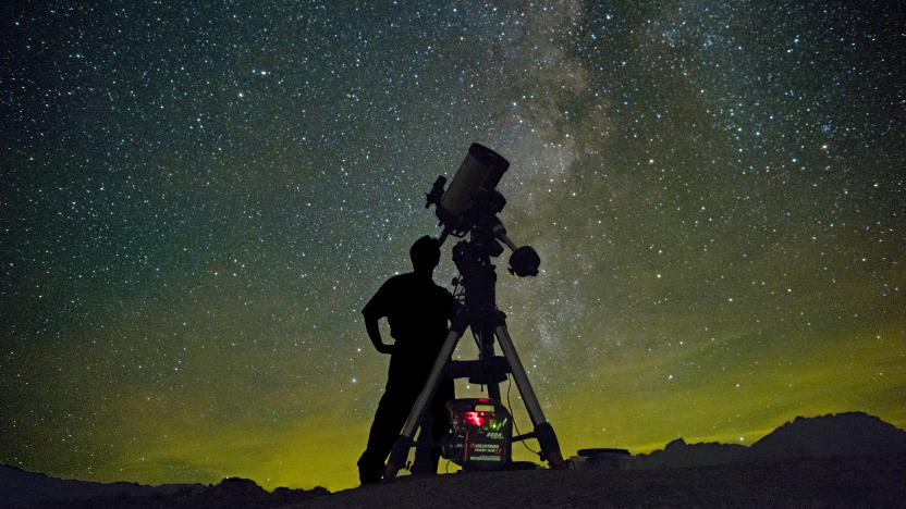An Astronomer with a telescope stands in silhouette against a starry night sky with a greenish yellow glow at the horizon line.