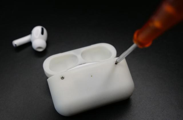 A custom AirPods Pro case with screws. A screwdriver is unscrewing one of the two visible screws. A lone AirPods Pro bud sits on the dark surface behind it.