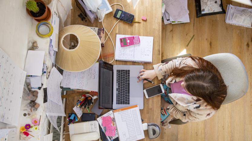 An overhead shot of a teenage Caucasian girl sitting at a desk at home studying. She is using a laptop and her desk is cluttered with books and such like.
