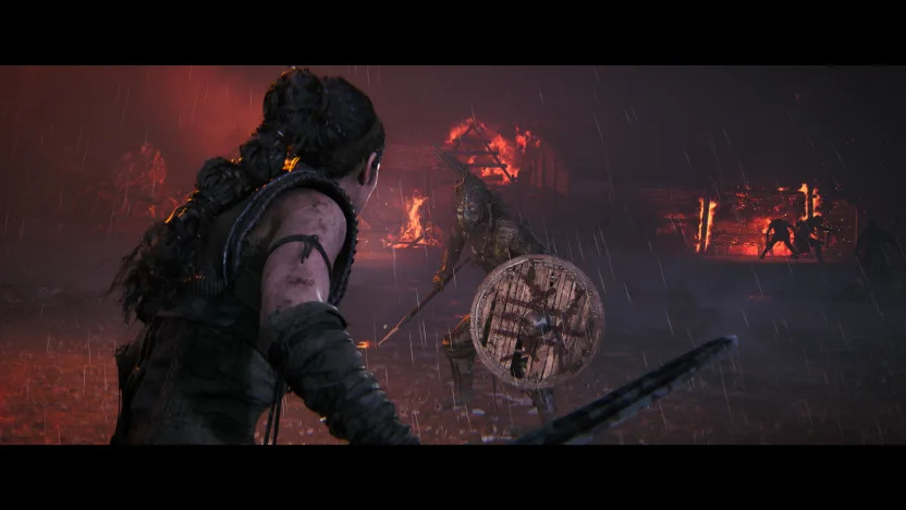 Dark setting, with the protagonist Senua wielding a sword in the foreground. A grotesque enemy with a shield lies ahead with burning buildings in the background.