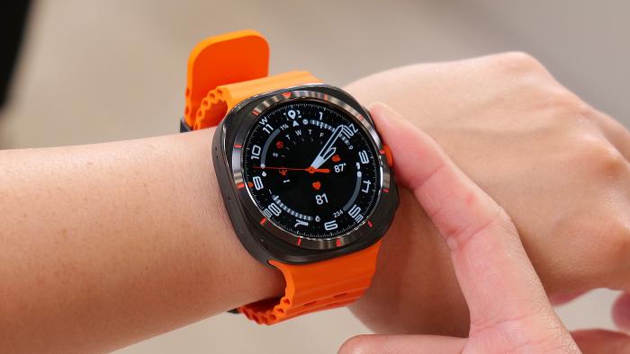 The Samsung Galaxy Watch Ultra on a wrist in mid air, showing an analog watch face with a black background and white and orange markings. A finger is resting against the middle button on the right edge.