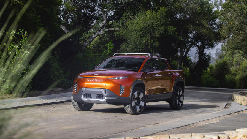 Image of an orange Fisker Alaska flatbed EV driving down a quiet road in front of some dark green foliage.