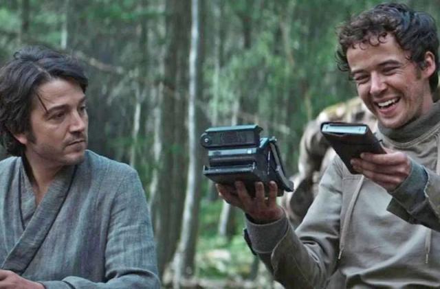 A still image from the tv series 'Andor' showing the main character sitting next to someone who's holding a device that appears to look like a folding polaroid camera. 