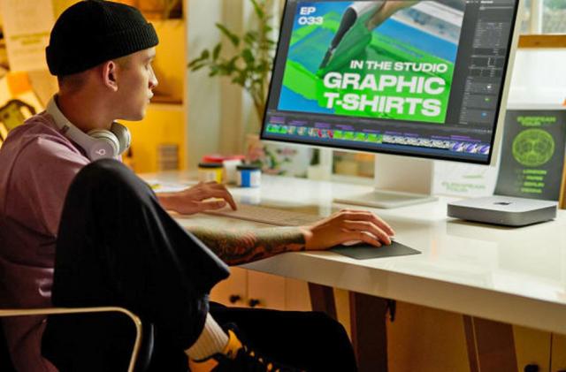 A graphic designer sits at their desk with a Mac Mini clearly visible on the desk top.