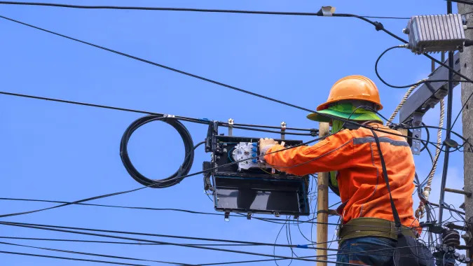 Rear view of technician on wooden ladder is working to install fiber optic and splitter box on electric pole against blue sky background