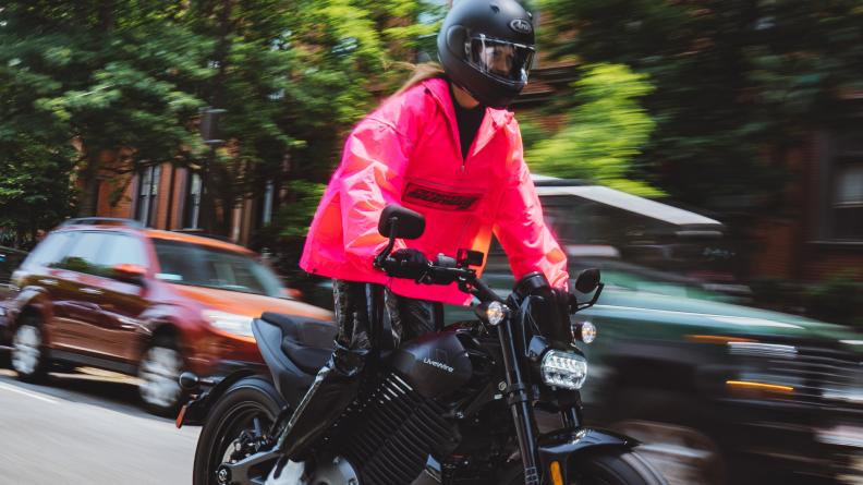 A person in a flamingo-pink windbreaker riding the LiveWire S2 Del Mar electric motorcycle down a city street. Cars are parked behind in front of trees and visible sections of city buildings.