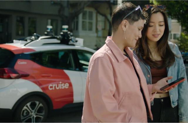 Two persons in the foreground, both looking down on a smartphone. In the background is a Cruise robotaxi painted red and white.