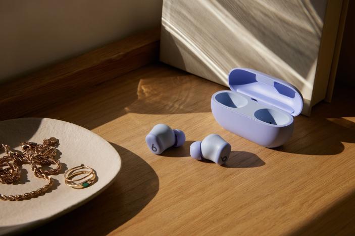 Beats Solo Buds in purple with both earbuds sitting on a wooden shelf outside of their case. A jewelry dish with rings, and other gold items sits in the foreground. 