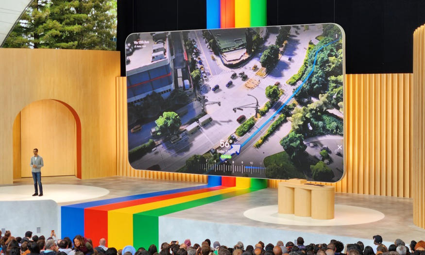 Photo of the stage at Google I/O keynote. Alphabet CEO Sundar Pichai is standing off to the left. A large screen shows a new feature in Google Maps, as 3D render of the route that the app is suggesting to a user. It shows a visualization of a city environment with a blue line and an arrow along a sidewalk. It shows the level of traffic, the temperature and the time of day.