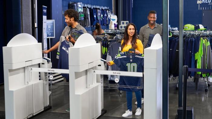 Customers use Amazon’s Just Walk Out tech in a merch store at Seattle’s Lumen Field. Three customers walk smiling as they prepare to exit through turnstiles.