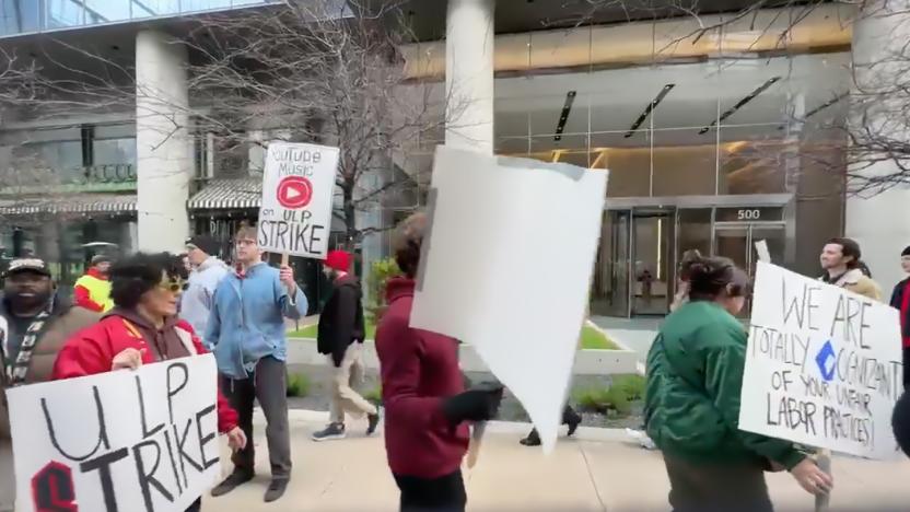 A picket line of strikers protesting outside of Google’s Austin, TX offices. Various people marching outdoors, holding picket signs demanding better treatment.