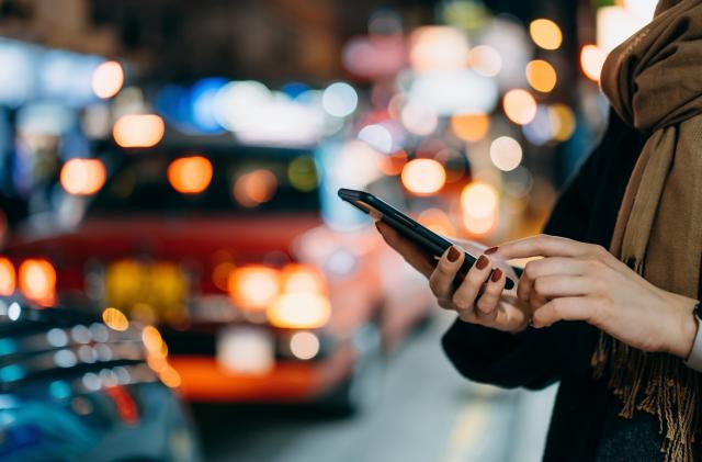 Close up of young woman using mobile app device on smartphone to arrange taxi ride in downtown city street, with illuminated busy city traffic scene during rush hour with traffic congestion in the evening