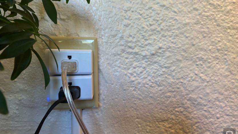Two smart plugs are in an outlet on a white wall. A white cord and a black cord are plugged in. A houseplant sits nearby.  