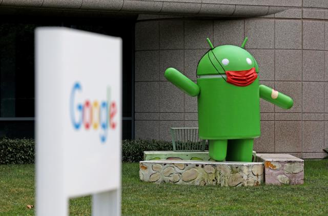 An image from outside of the Google campus building in Mountain View, CA. showing an out of focus Google sign in the foreground and an Android statue with an ad-hoc face mask behind it.

