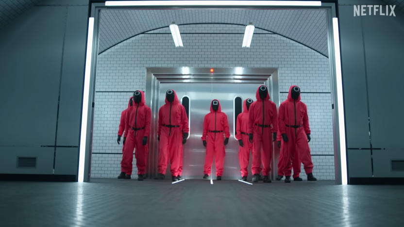 People dressed as Squid Game guards (reddish-pink suits with black faces) stand in a cold industrial building.