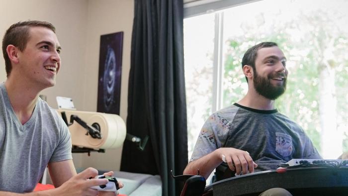 Xbox marketing photo showing two young men playing Xbox games. One of them is using a standard Xbox gamepad, and the other has the Xbox Adaptive Controller.