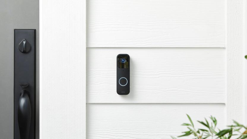 Marketing photo of a Blink Video Doorbell (horizontally oblong device, black, with a camera sensor on top and blue-ring button on bottom) on a white panel outdoor wall next to a front door.