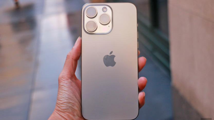 The iPhone 15 Pro Max held in mid-air with a sidewalk in the background.