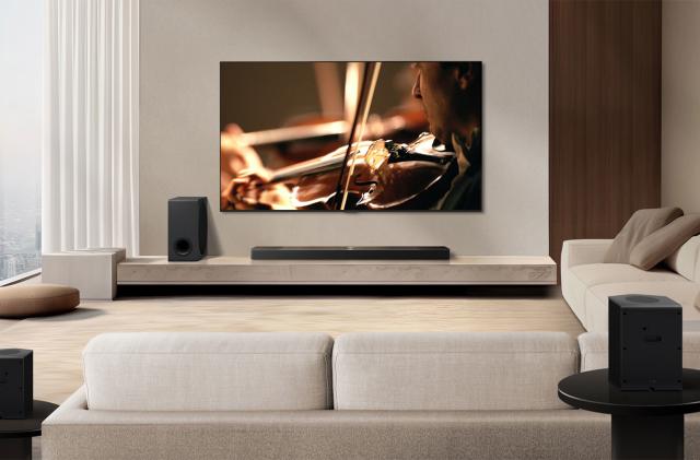 A living room with a black soundbar and subwoofer underneath a flatscreen TV. An angled sofa is in the frame with two rear speakers behind one side of it. 