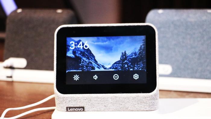 A gray Lenovo Smart Clock 2 on a wireless charging dock with its screen facing the camera. A row of options for volume, brightness, do not disturb and settings take up the bottom of the display.  In the background are a black and a blue Smart Clock 2 on the left and right respectively.
