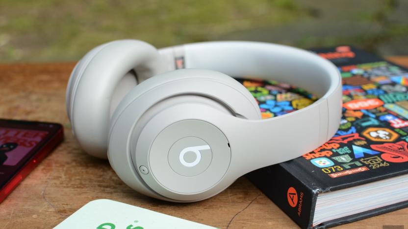 A white pair of Beats Studio Pro over-ear headphones sit on a wooden tabletop as it leans on a hardcover book.