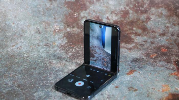 Photo of the Samsung Z Flip 5 sitting on a surface that looks like rough metal with rusty spots. The phone is partially open with its camera view visible on the top half of the screen.