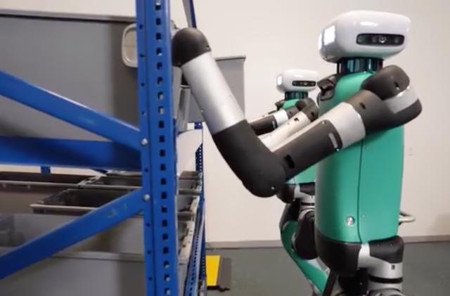 Two humanoid bipedal robots reaching onto warehouse shelves to grasp plastic storage bins.