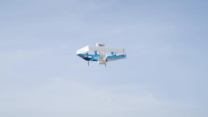 Amazon marketing photo of a Prime Air drone (mostly white with blue accents) flying in the sky. Tops of trees visible below.
