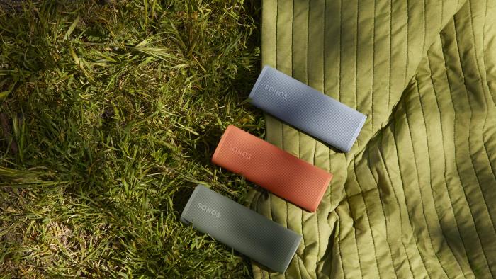 Three Sonos Roam 2 portable speakers (green, red, blue), sitting side-by-side (diagonal to the camera) on an outdoor blanket on top of grass.