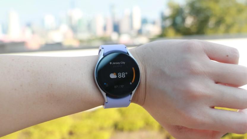 Photo of an arm wearing the Samsung Galaxy Watch 5 with lavender band against a background of trees, grass and buildings.