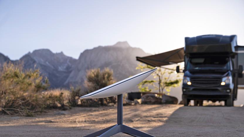 A Starlink satellite dish sits on a four-legged stand in a desert-style environment next to a black RV.