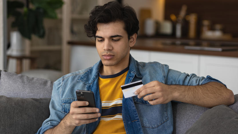 Millennial european man holding plastic credit card in hands, makes purchases on the Internet or uses online banking services sitting on the couch at home