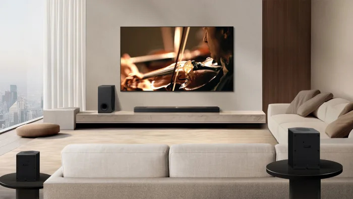 A living room with a black soundbar and subwoofer underneath a flatscreen TV. An angled sofa is in the frame with two rear speakers behind one side of it. 