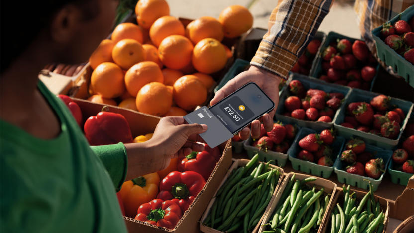 A person wearing a green shirt holding a credit card over an iPhone to pay for their purchase. Oranges, bell peppers, strawberries and string beans in containers are in the  background.