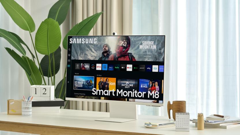 A press shot displaying the Samsung Smart Monitor M8 on a light brown wooden table next to a potted houseplant and an assortment of desktop accessories.