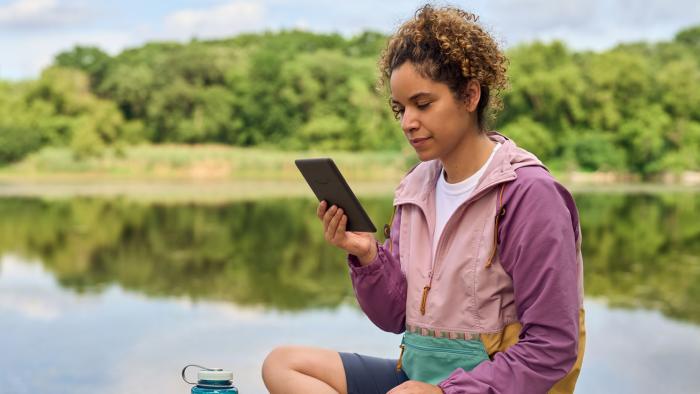 A woman sits on a rock near a lake reading from an Amazon Kindle