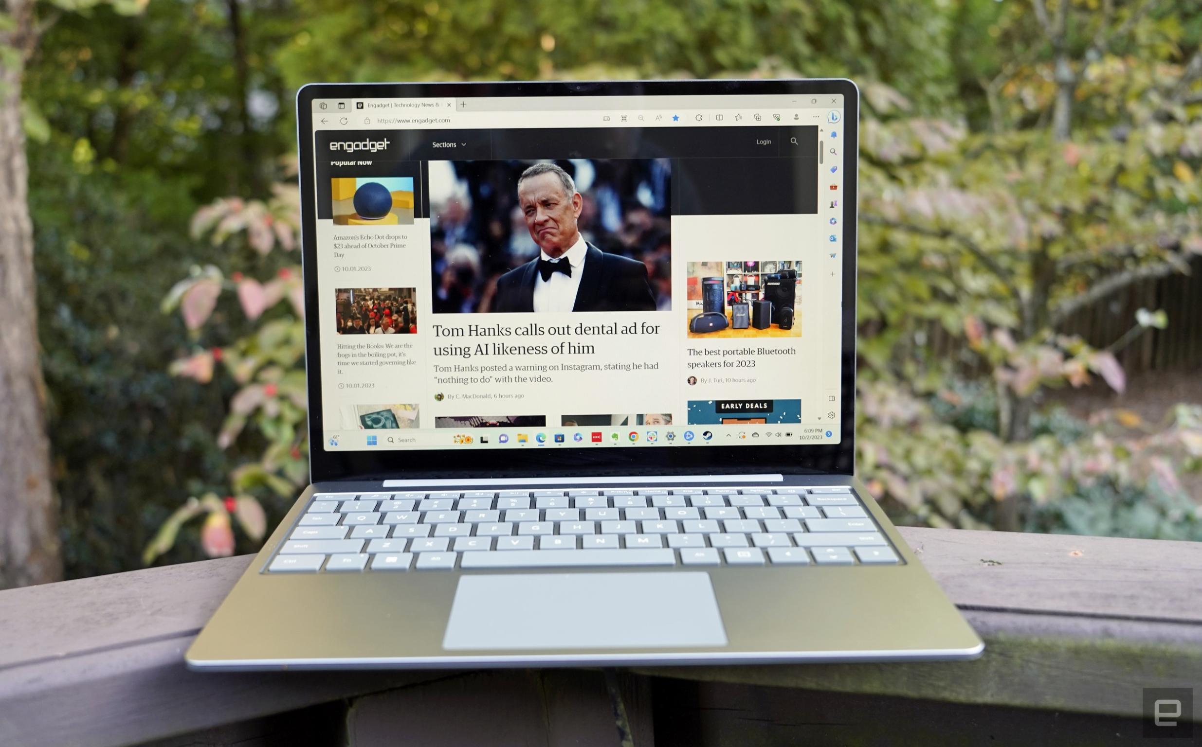 A Surface Laptop Go 3 sits open on a backyard deck railing with foliage in the background.