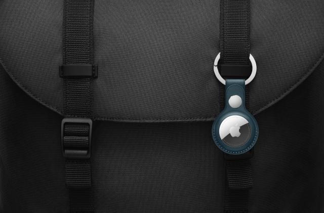 An Apple AirTag in a navy blue case clipped to the outside of a black backpack