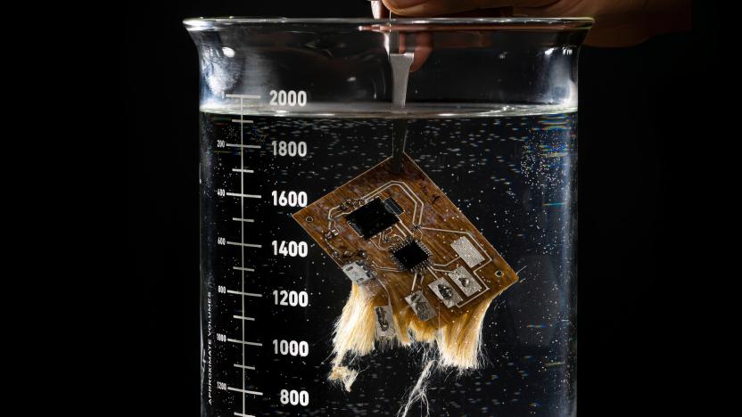 Photo of a printed circuit board (PCB) in a glass measuring cup, dissolving in water. Its fibers are coming apart near the bottom.