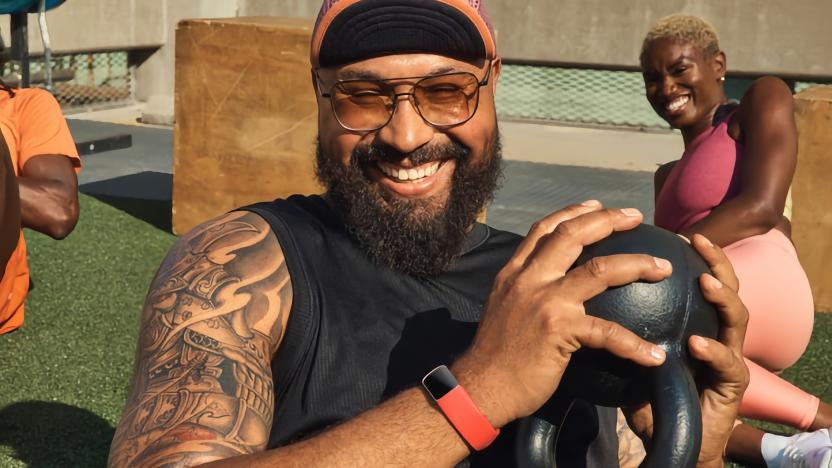 Lifestyle marketing photo of a man holding a small kettlebell in his hand with a (red band) Fitbit Charge 6 on his wrist. A woman sits behind him, stretching, as the two laugh together in an outdoor fitness area.