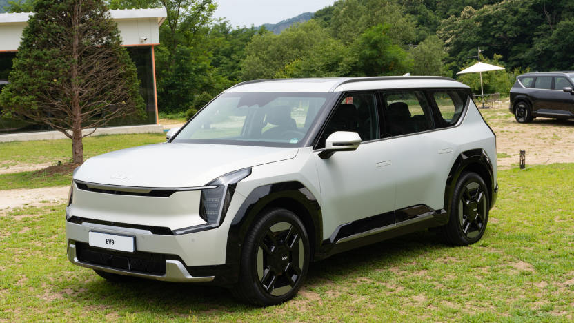 A white Kia EV9 SUV is parked in a grassy park area with woods in the background.