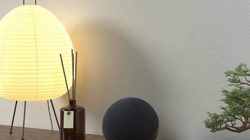 An Echo Show sits on a wooden table beside a diffuser and lamp.