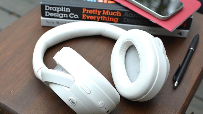 White headphones sitting on a wooden table from above with a red notebook and phone in the background sitting on top of a black and a white book.