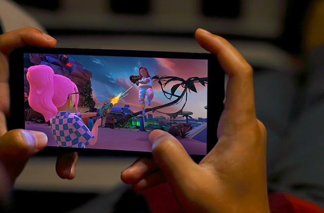 Hands holding a phone with a game on screen. The game shows a pink-haired character shooting at another character in the background.