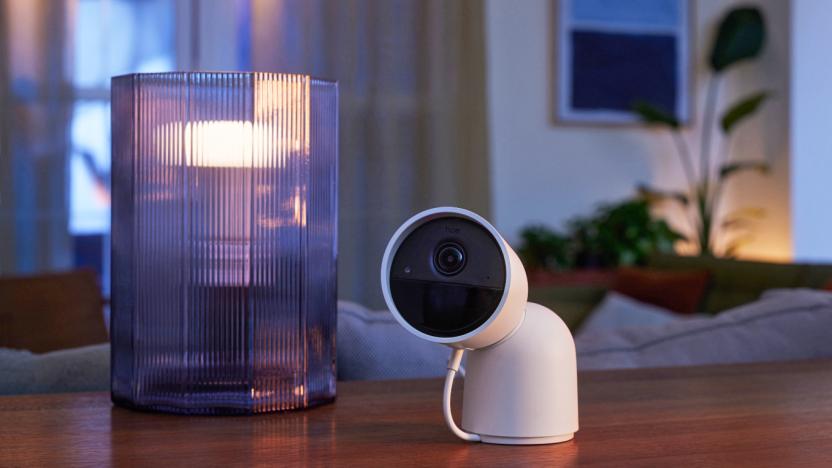 A white Philips Hue Secure camera with an articulated head is tilted towards the camera while it sits on a wooden dining room table next to a light that's in a fluted glass jar.