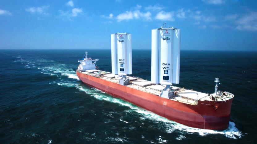 A cargo ship fitted with two giant (white-colored) rigid sails floats on the open sea. The enormous ship has reddish sides and white-ish deck.