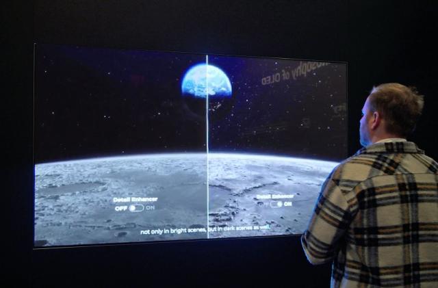 A man stands in front of a large TV display in a dark room. The screen's image is split in half (left and right) to show the differences of picture quality.