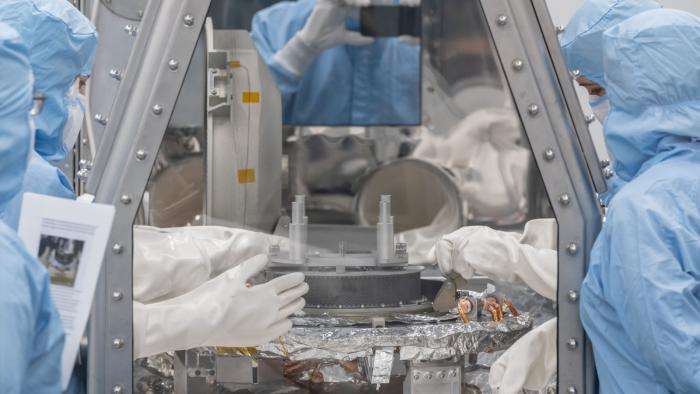 Scientists in blue overalls standing around a glass container with their gloved hands inside.